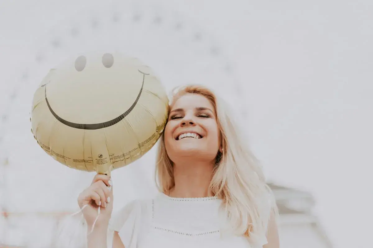 Uśmiechnięta blondynka trzymająca balon z buźką, jakby usłyszała świetny żart po angielsku.