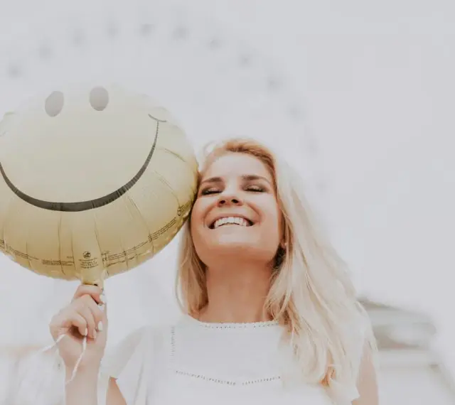 Uśmiechnięta blondynka trzymająca balon z buźką, jakby usłyszała świetny żart po angielsku.
