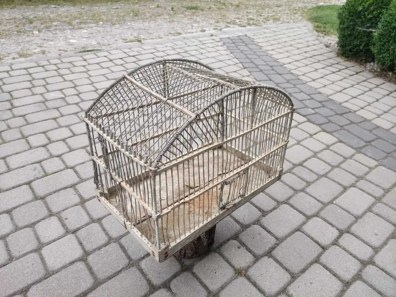 Stara, metalowa klatka dla kanarka retro, z drewnianą podstawą, stoi na wybrukowanym podwórku.